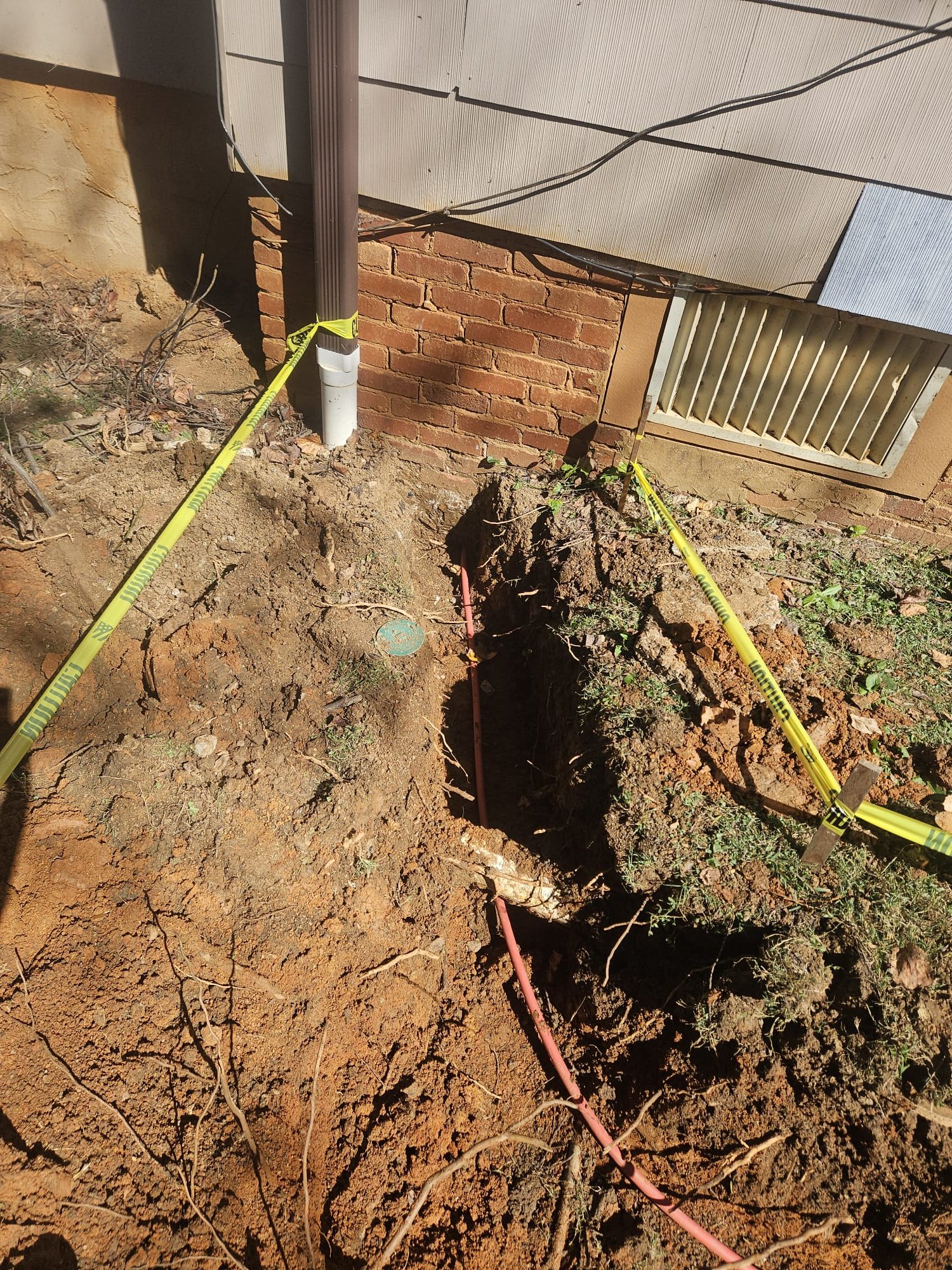 Outdoor trench with new red pipe connected to downspout and caution tape