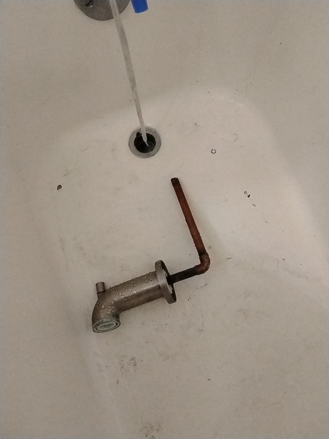 This image shows the inside of a bathtub. Water is running from a pipe directly into the drain, and there is an old, removed bathtub spout with a copper pipe attached lying in the tub. The spout and pipe appear to be used and show signs of age, such as water spots and some corrosion on the copper. The tub itself has some dirt and marks, likely from the removal process.