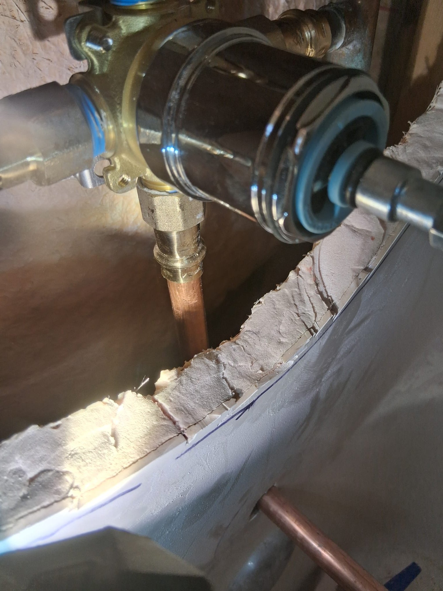 This image shows a close-up view of plumbing work inside a wall, likely related to a shower or bathtub installation. The main focus is on a new brass and chrome valve assembly, which appears to be recently installed. The copper pipes connected to the valve are also new and clean, indicating recent plumbing work.